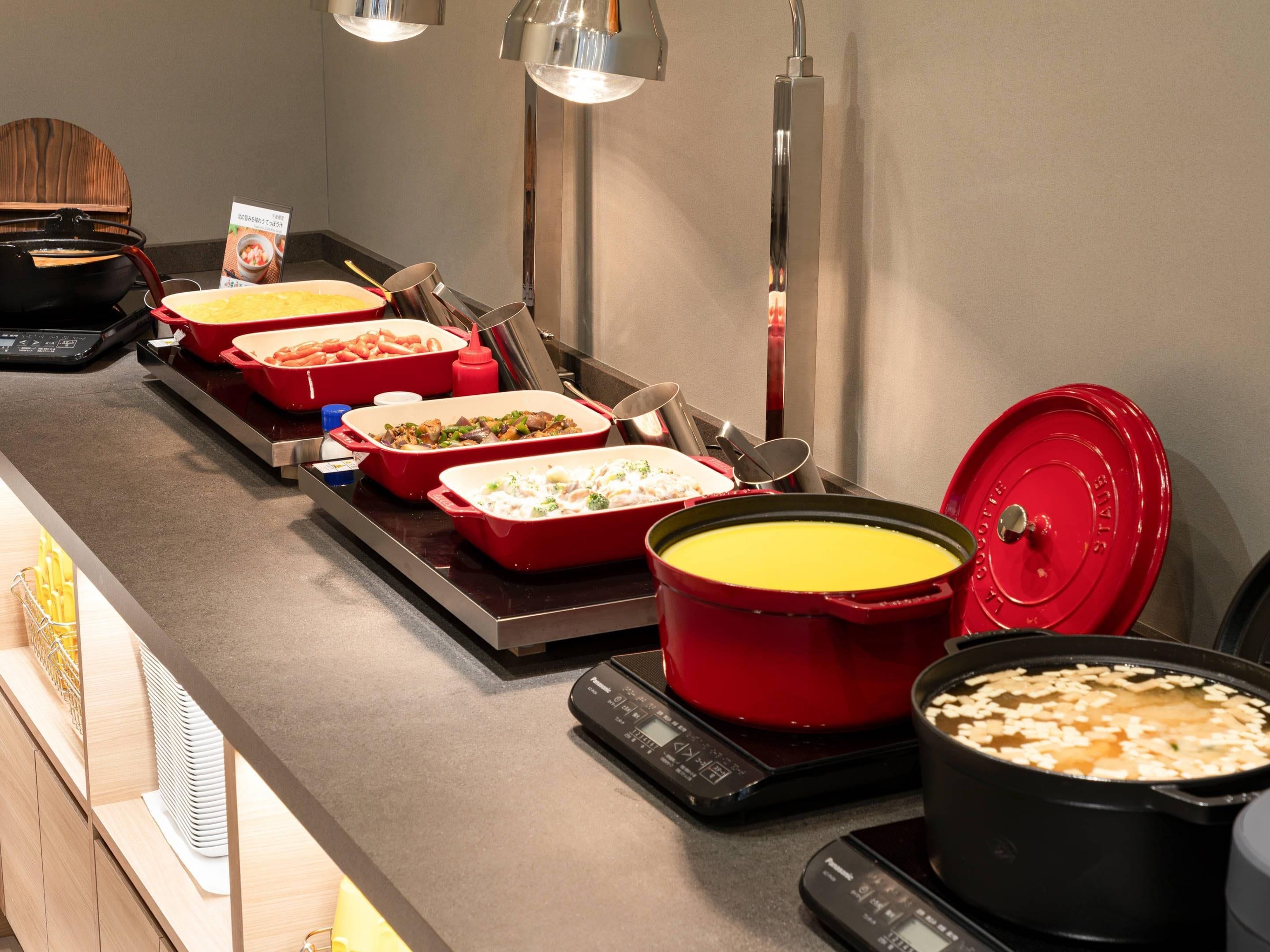 A buffet setup with various dishes in red and black containers, a weighing scale, and a large pot of soup.