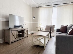 Un soggiorno moderno con una TV su un mobile in legno, un tavolino da caffè bianco, un divano grigio e una lampada da terra.