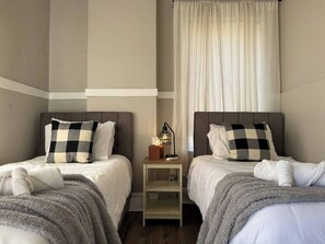 Una camera d'albergo con due letti, ognuno con un cuscino a quadretti, un comodino con una lampada e una finestra con tende.
