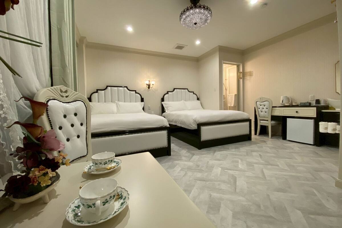 Habitación de hotel con dos camas, un escritorio, una silla, un refrigerador pequeño y un candelabro decorativo.