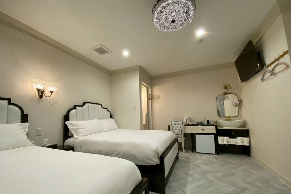 Habitación de hotel con dos camas, un escritorio con espejo, un lavamanos pequeño y un televisor en la pared.