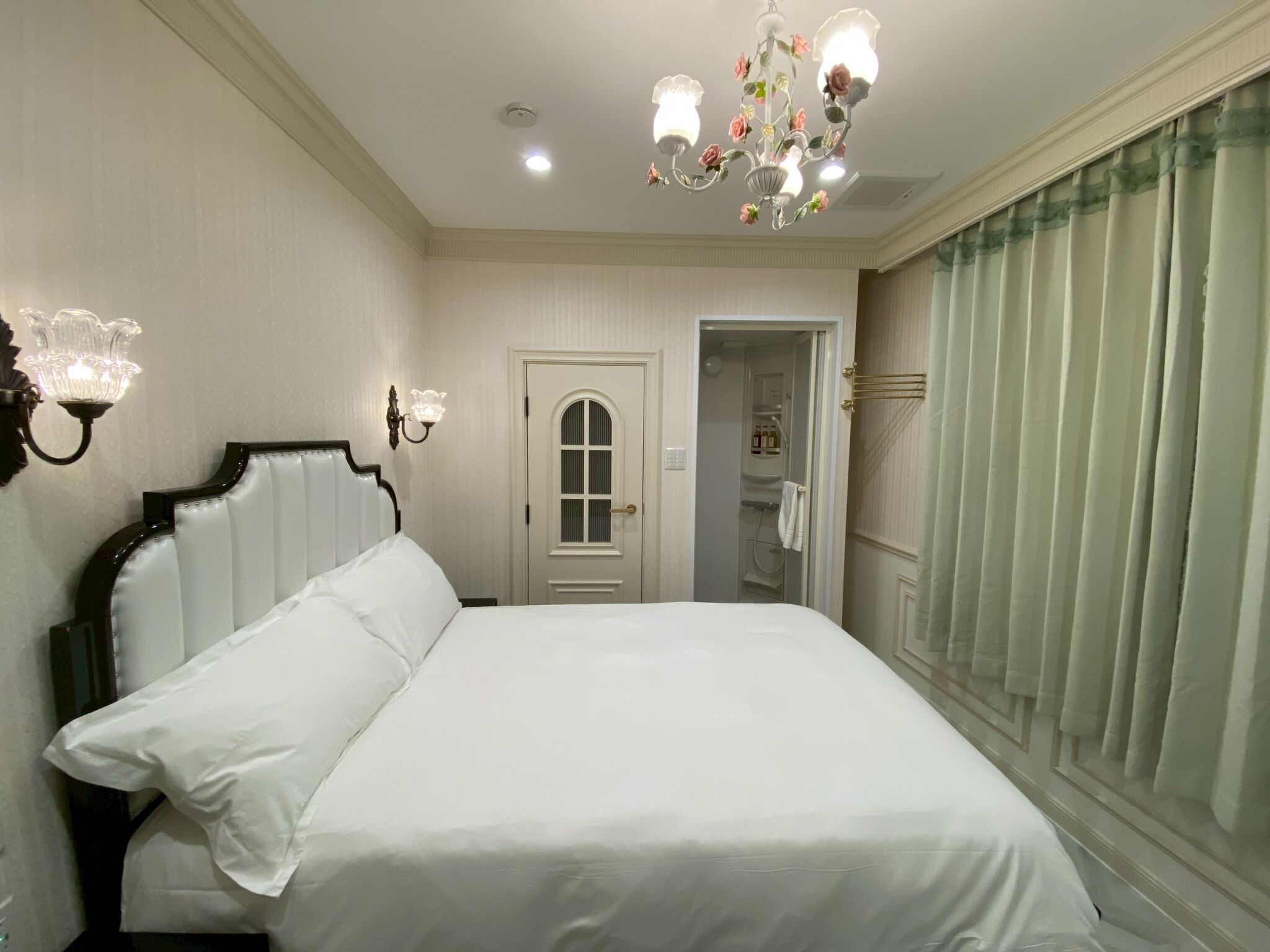 Un dormitorio ordenado con una cama grande, una araña de cristal, una puerta que da al baño y una ventana con cortinas.