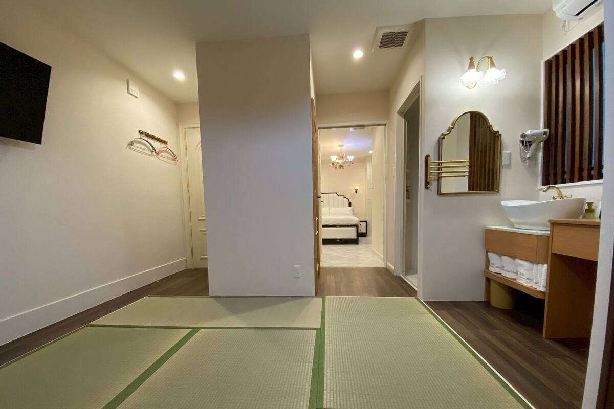 Habitación de hotel con cama, baño con lavamanos y espejo, y un pasillo que conduce a otra habitación.