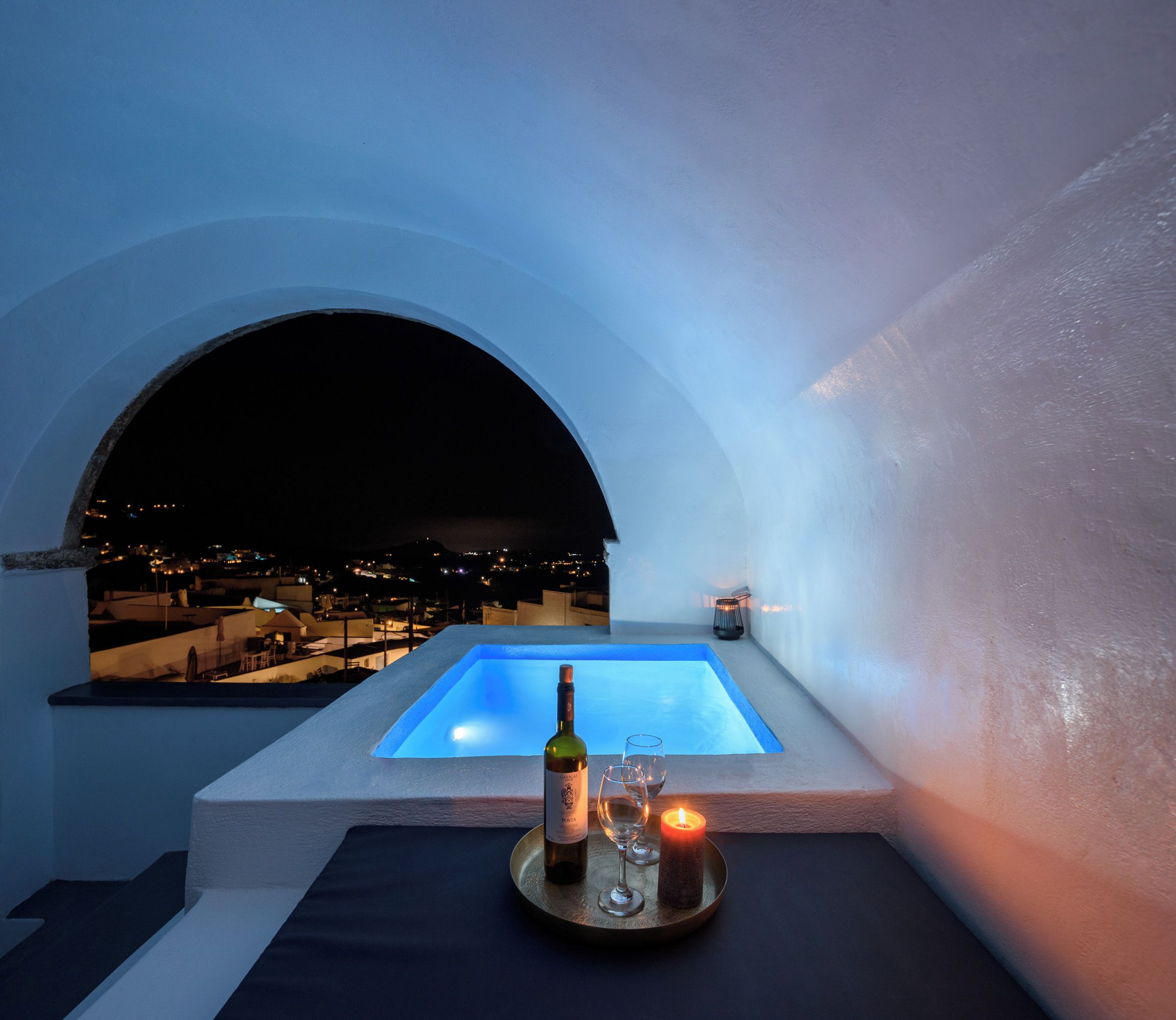 A rooftop terrace with a pool, a bottle of wine, and glasses on a tray, overlooking a cityscape at night.