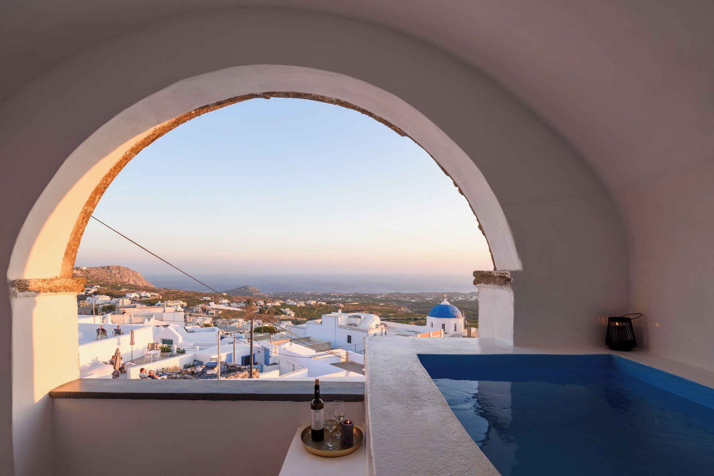 Vista desde una habitación con un ventanal arqueado blanco que da a un pueblo costero con edificios blancos y cúpulas azules.