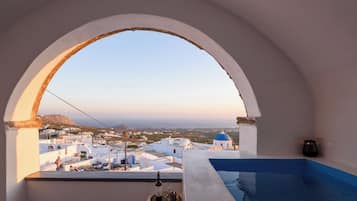 Vista desde una habitación con un ventanal arqueado blanco que da a un pueblo costero con edificios blancos y cúpulas azules.