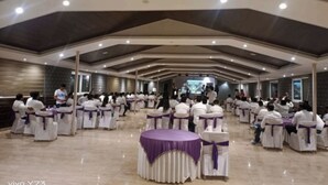 Un salón amplio con filas de sillas y mesas blancas, algunas con cintas moradas, bajo un techo alto con iluminación empotrada.