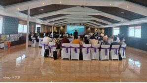 Un salón de conferencias con un escenario al frente, sillas blancas adornadas con cintas moradas y un suelo de madera pulida.