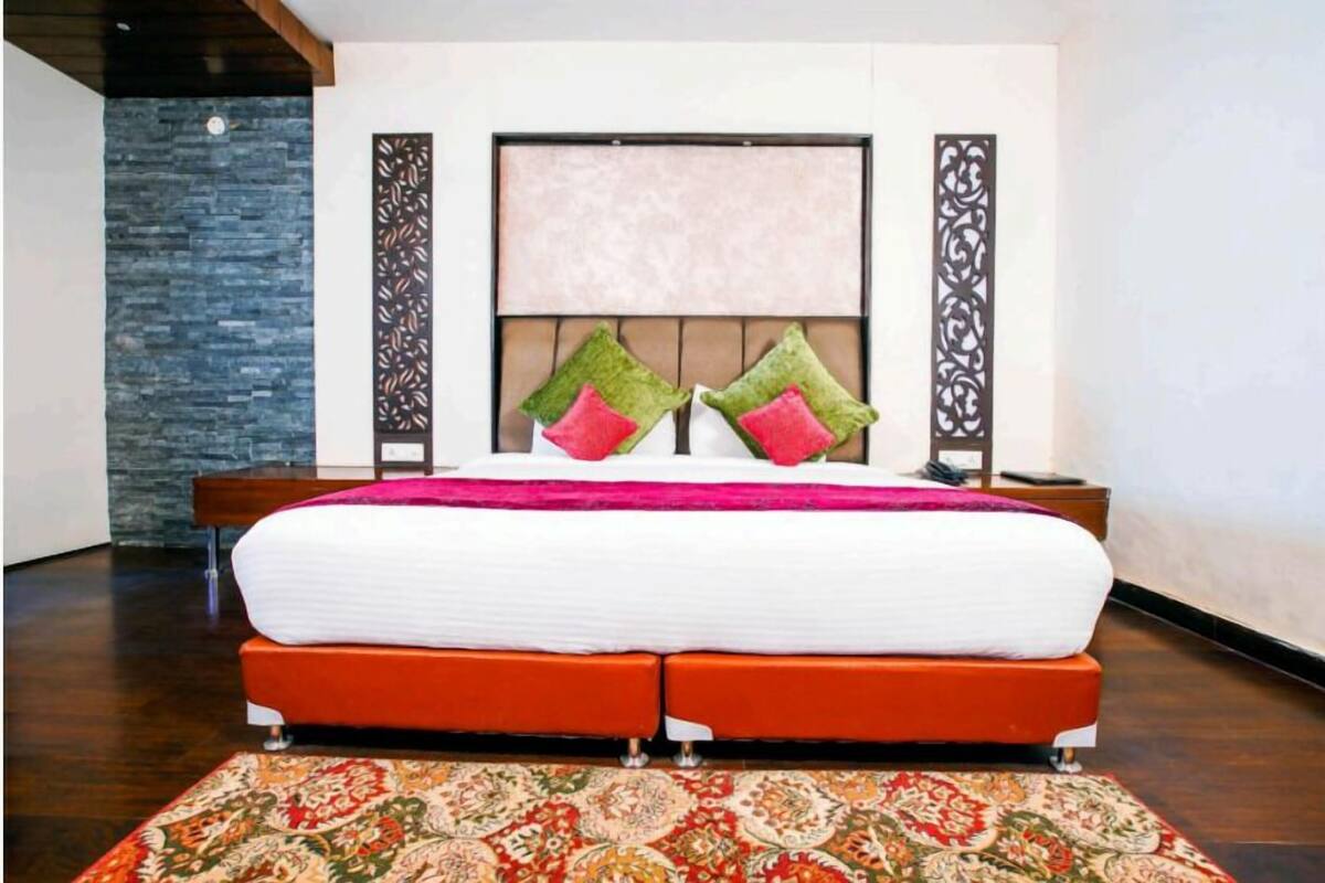 Cama doble con colchón blanco y tablero de cabecera naranja, flanqueada por dos almohadas decorativas, una manta rosa y una alfombra estampada.