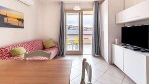 Ein modernes Zimmer mit einem Esstisch, einer Couch mit gemusterten Kissen, einem Fernseher auf einer Kommode und einem großen Fenster mit Vorhängen.