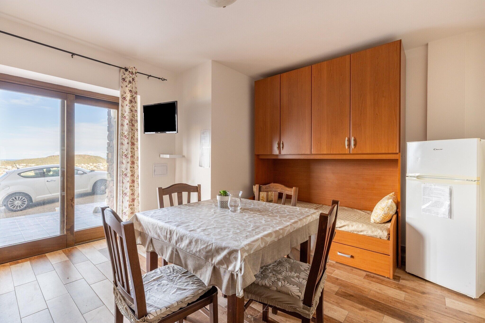 A compact kitchen with wooden cabinets, a refrigerator, a dining table with chairs, and a sliding glass door leading to a parking area.