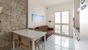 A room with a stone wall, a painting, a sofa, a dining table with chairs, and a kitchen area. - Beachfront terrace and air conditioning (Castelsardo)