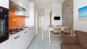 A modern kitchen with white cabinets, an orange backsplash, and a dining area with a stone wall and a painting of a beach. - Beachfront terrace and air conditioning (Castelsardo)
