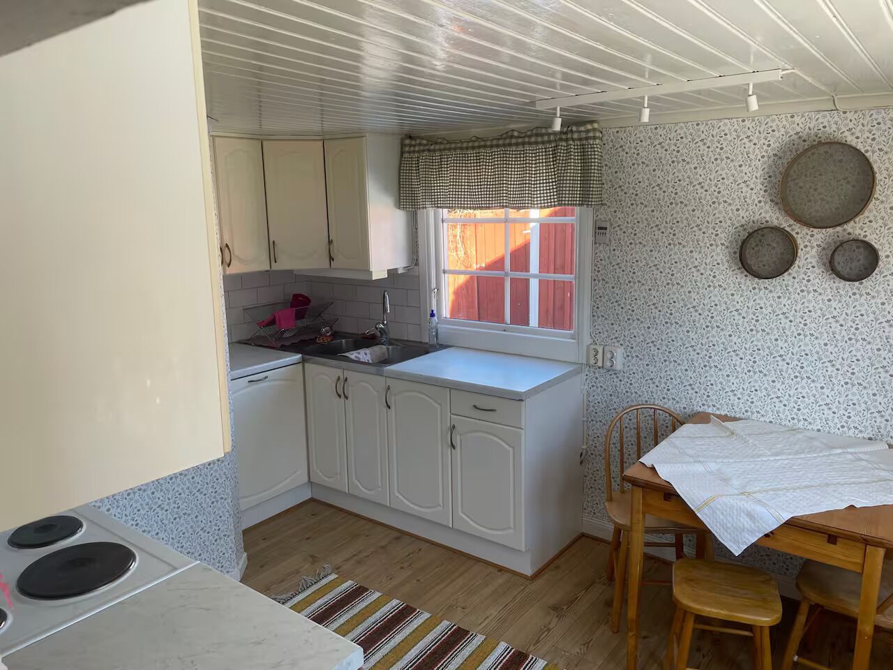 Eine kompakte Küche mit weißen Schränken, einem Fenster mit kariertem Vorhang und einem Essbereich mit Holztisch und Stühlen.