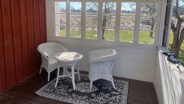 Une véranda abritée avec deux fauteuils en osier et une petite table, un tapis à motifs noir et blanc, et une vue sur un champ verdoyant par de grandes fenêtres.