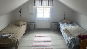 Une petite chambre mansardée avec deux lits simples, une fenêtre avec des rideaux à rayures, un radiateur et un plancher en bois.