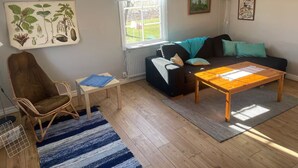Un salon avec un plancher en bois, un fauteuil marron, un canapé noir avec des coussins turquoise, une table basse en bois et une fenêtre avec des rideaux blancs.