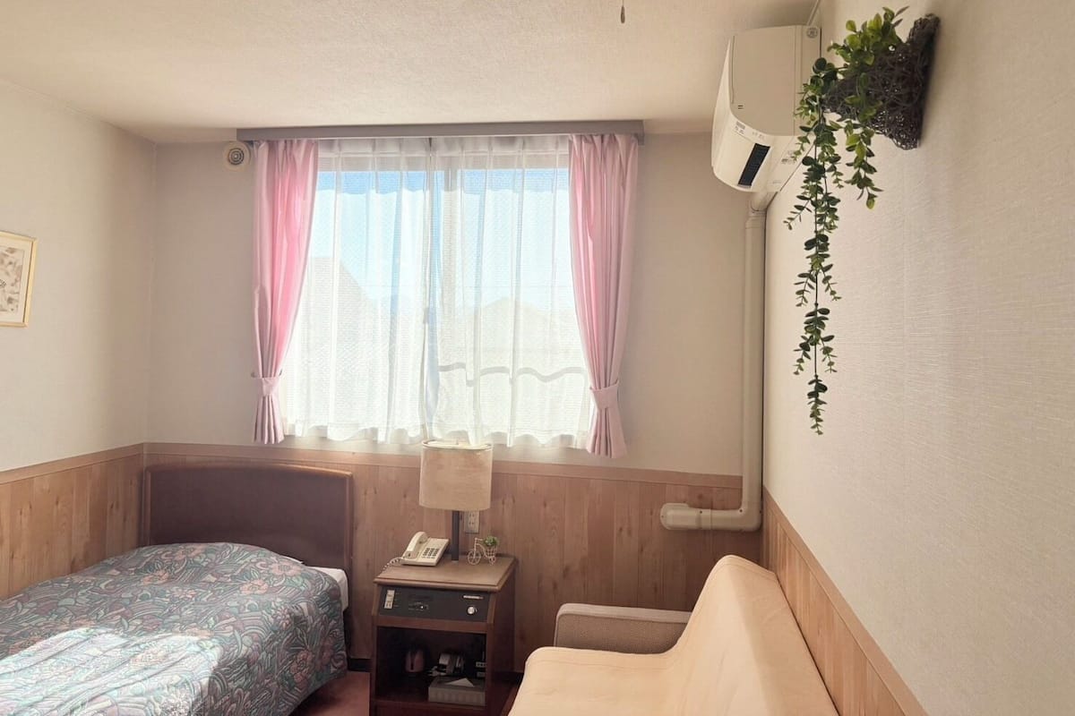 A hotel room with a bed, a small bedside table with a lamp, a chair, a window with sheer curtains, and an air conditioning unit.