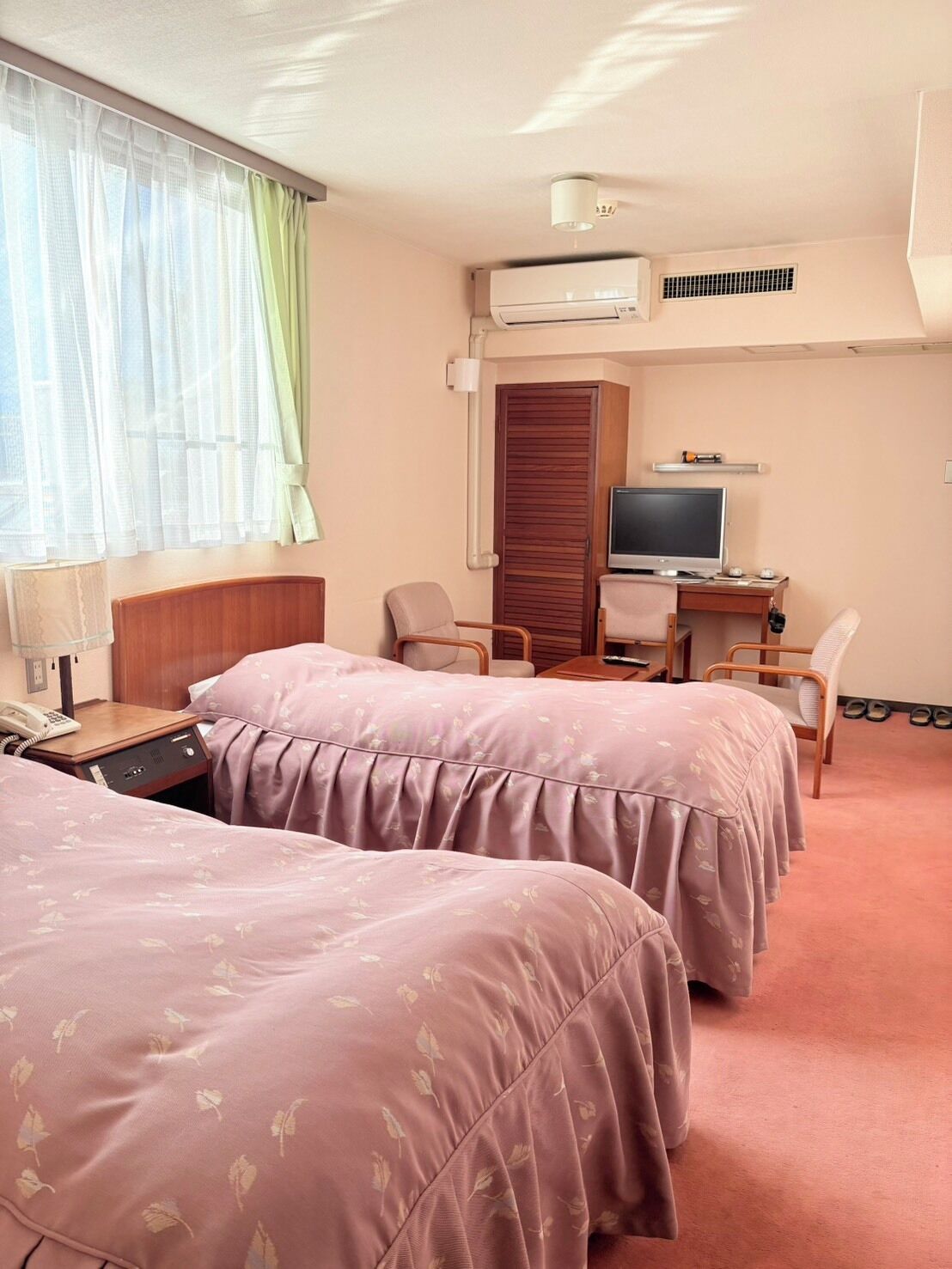 A hotel room with two beds, a desk with a television, a chair, a lamp, and an air conditioning unit.