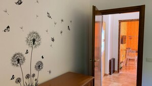 Une pièce avec une commode en bois, un mur décoré de pissenlits et de papillons, et une porte donnant sur un couloir avec des rideaux orange.