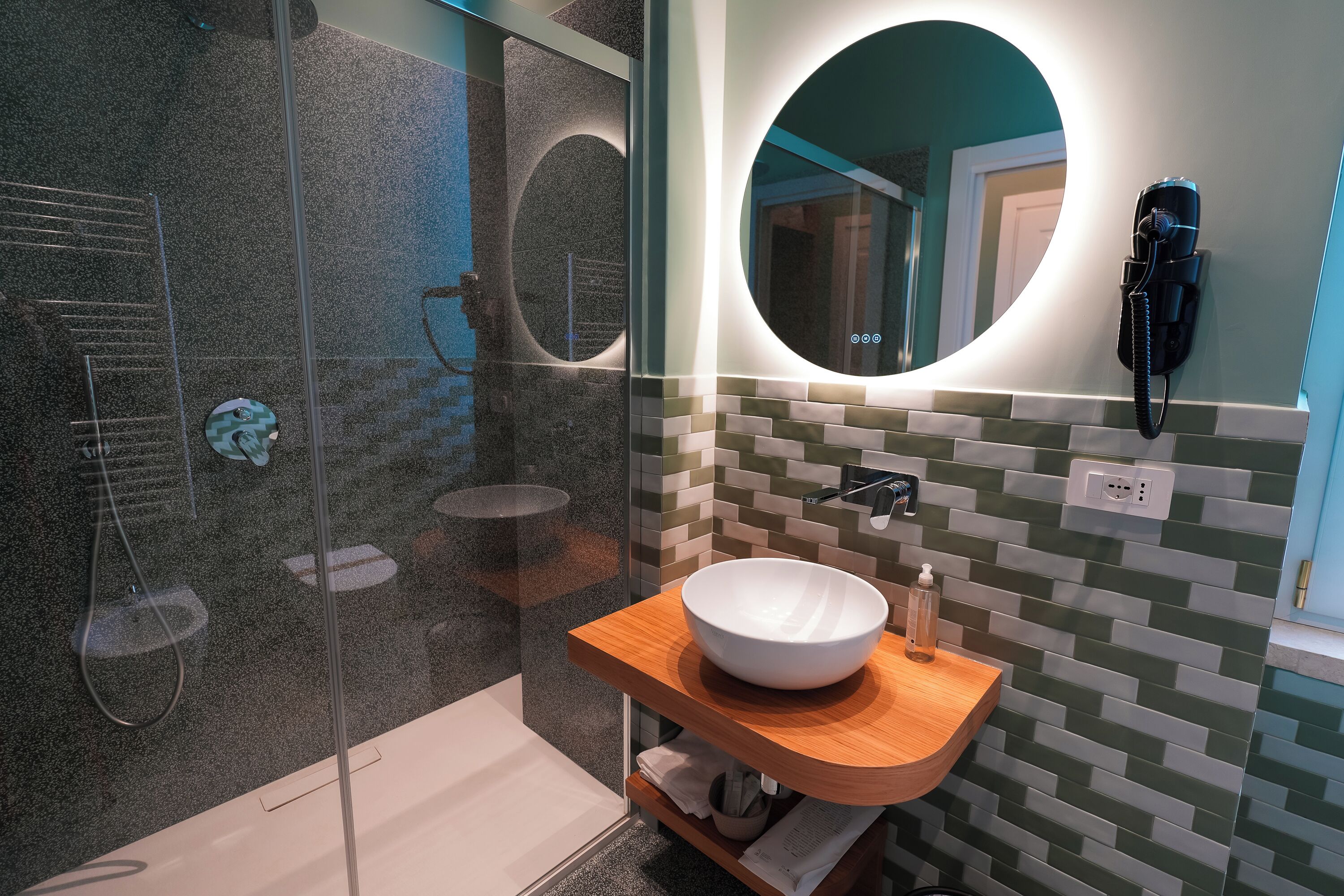 A modern bathroom with a glass shower enclosure, a wooden vanity with a white sink, and a round mirror with a built-in phone.