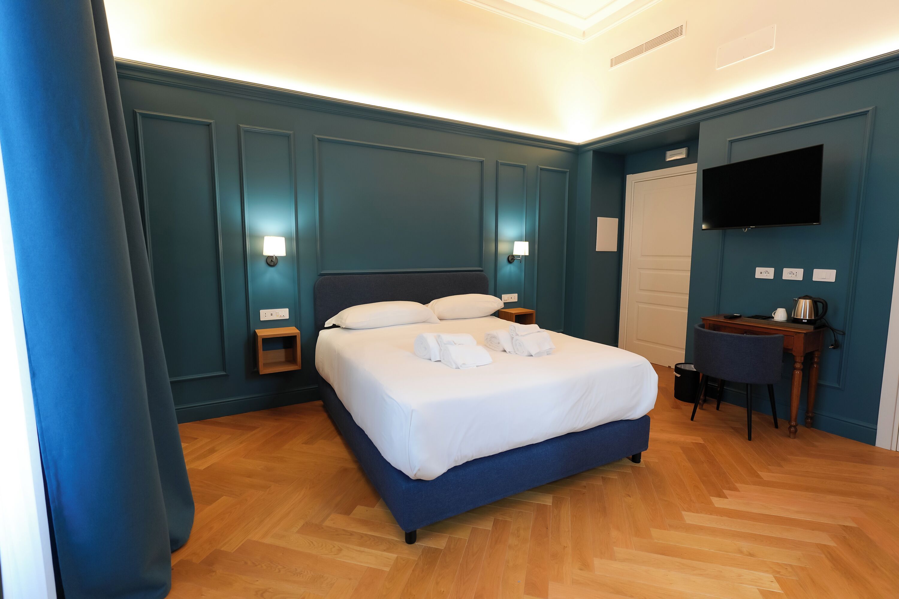 Un lit bien fait, avec du linge de lit et des serviettes blanches, une table de chevet en bois, un bureau avec une chaise et un téléviseur à écran plat fixé au mur.