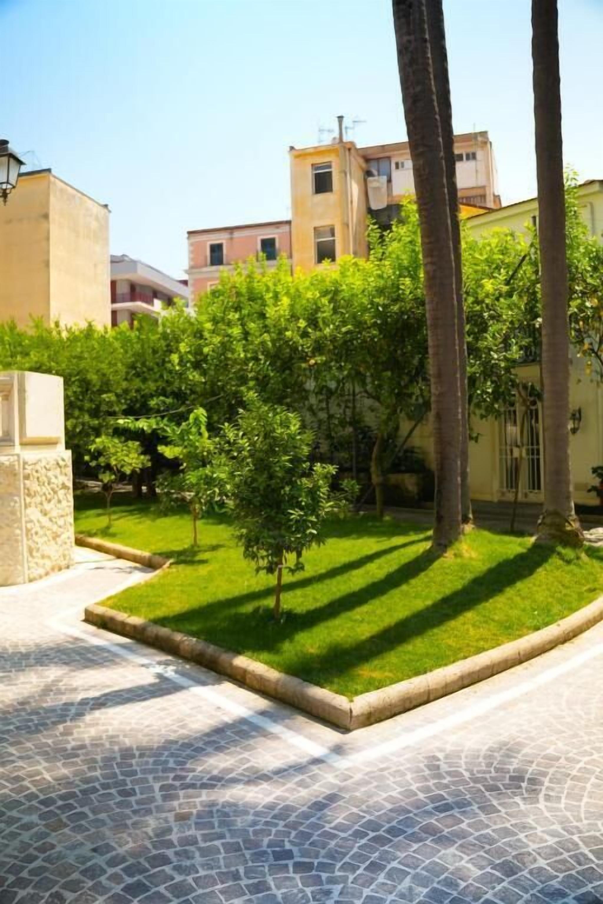 Un patio con un sendero empedrado, rodeado de follaje y palmeras, que conduce a un edificio con fachada color crema.