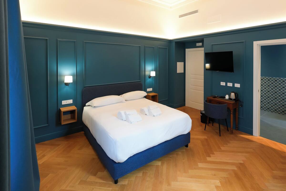 Une chambre d’hôtel bien aménagée, avec un grand lit, des lampes de chevet, une télévision et une petite table sur laquelle se trouve une bouilloire.