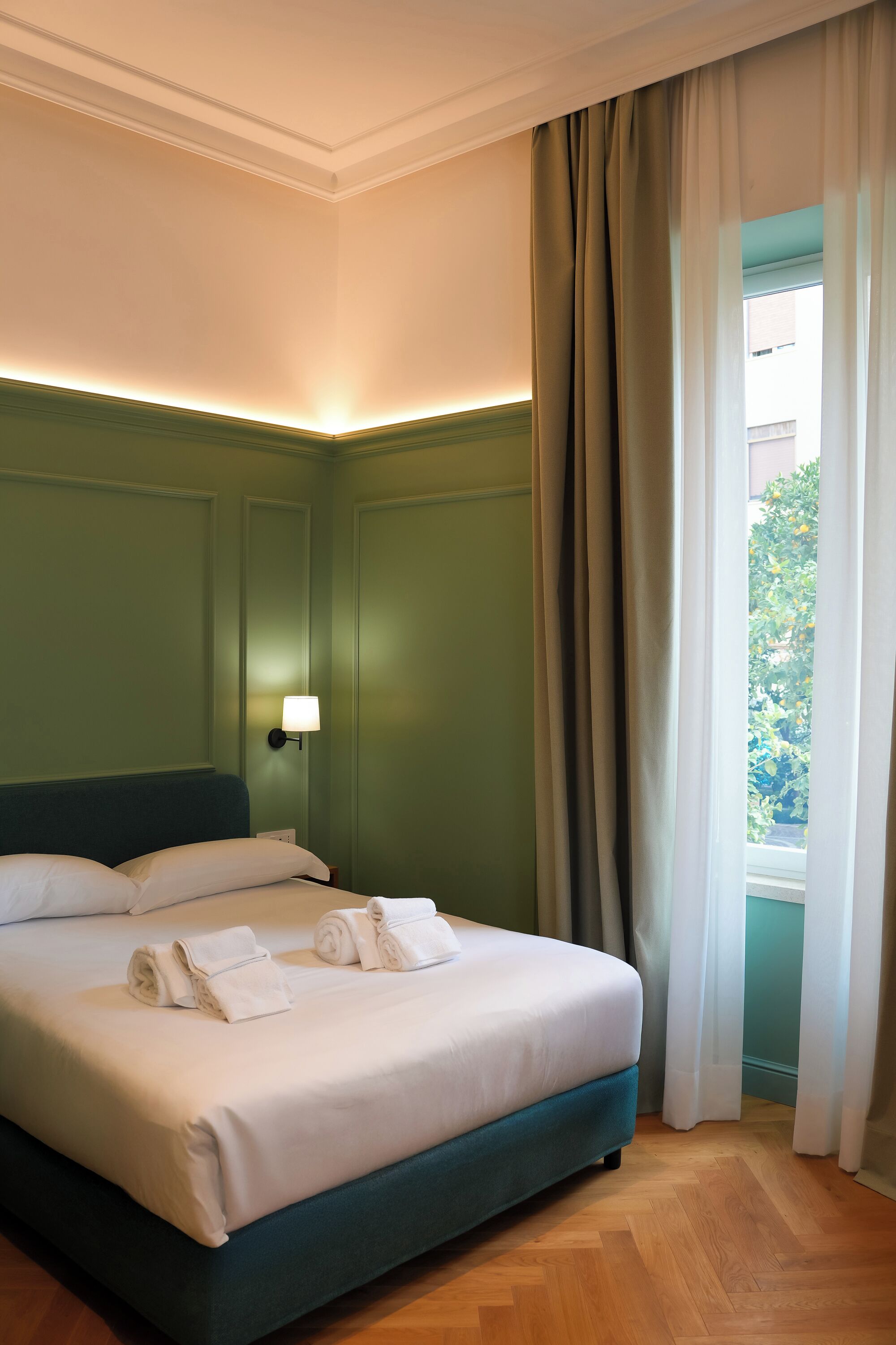 Un letto rifatto con lenzuola bianche e asciugamani arrotolati, una lampada a parete e vista sul verde attraverso una finestra con tende.