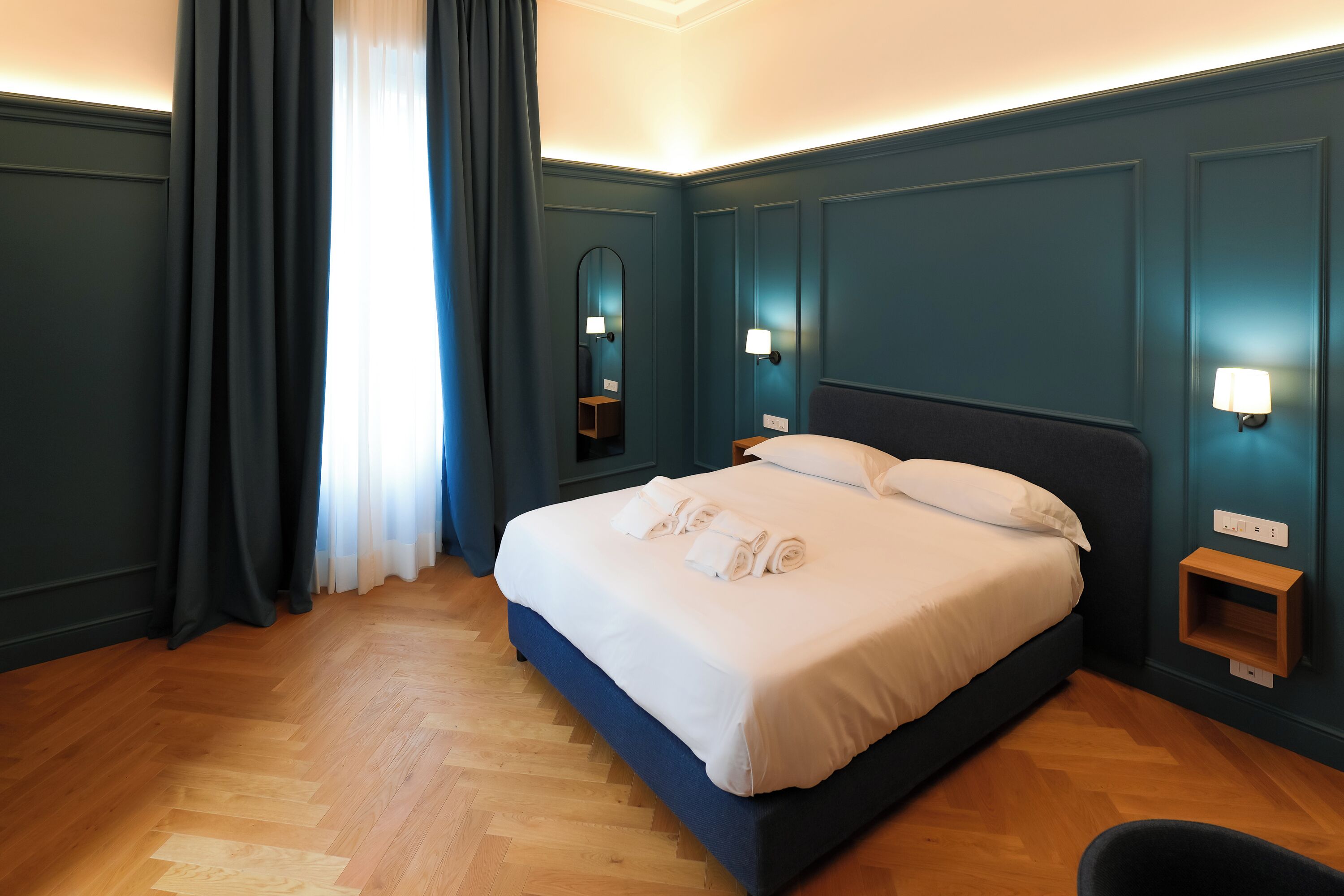 A neatly made bed with white linens and rolled towels, a wooden floor, and a wall-mounted nightstand with a lamp.