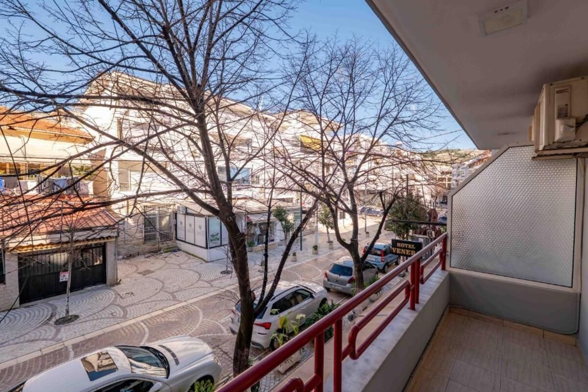 A balcony with a red railing overlooking a street with parked cars and buildings.