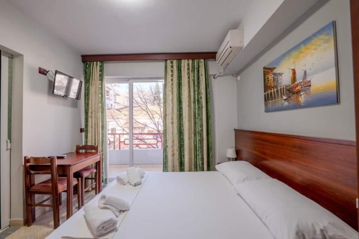 A hotel room with a bed, a desk with chairs, a TV, and a painting on the wall.