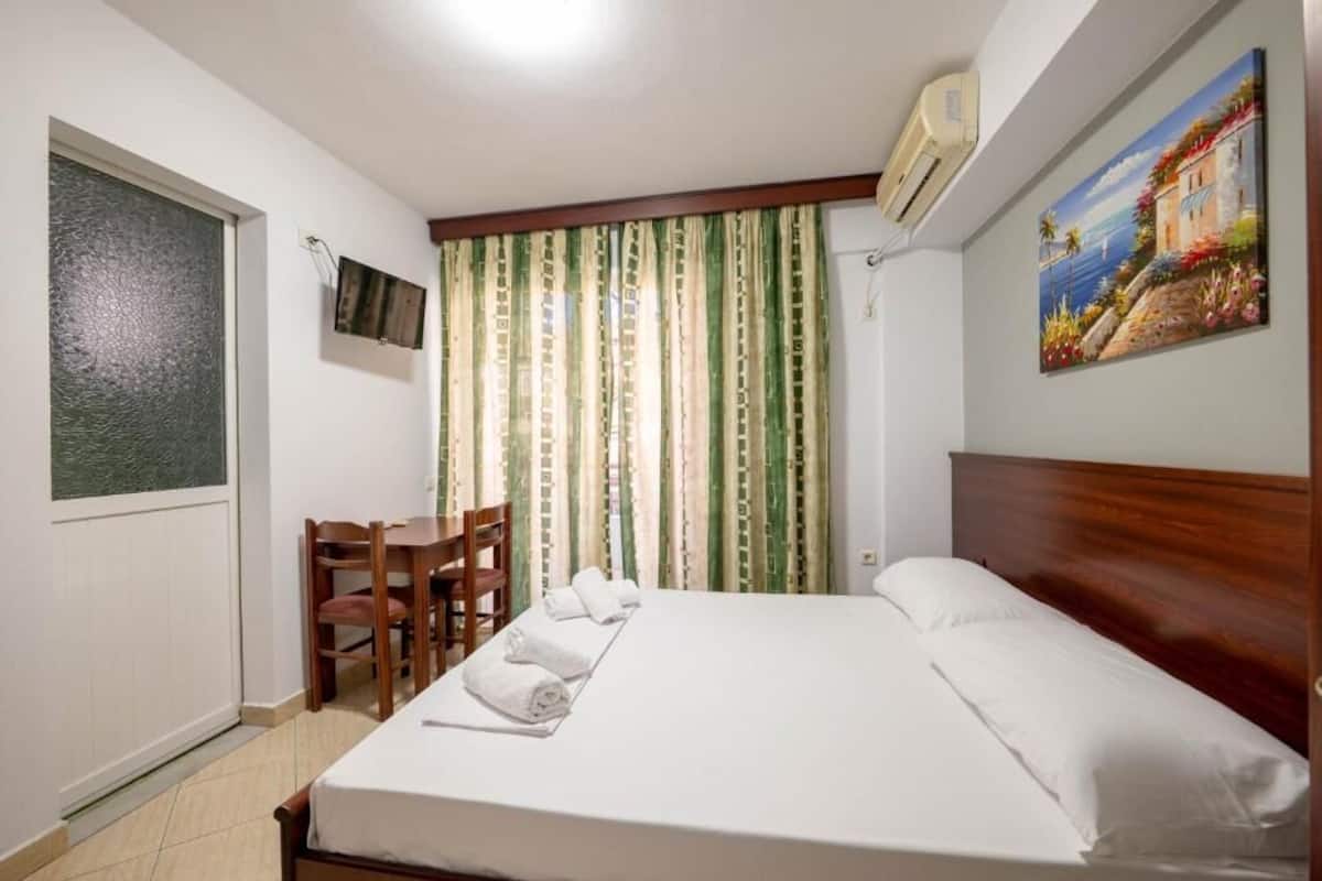 A hotel room with a bed, a desk with chairs, a television, and a painting on the wall.