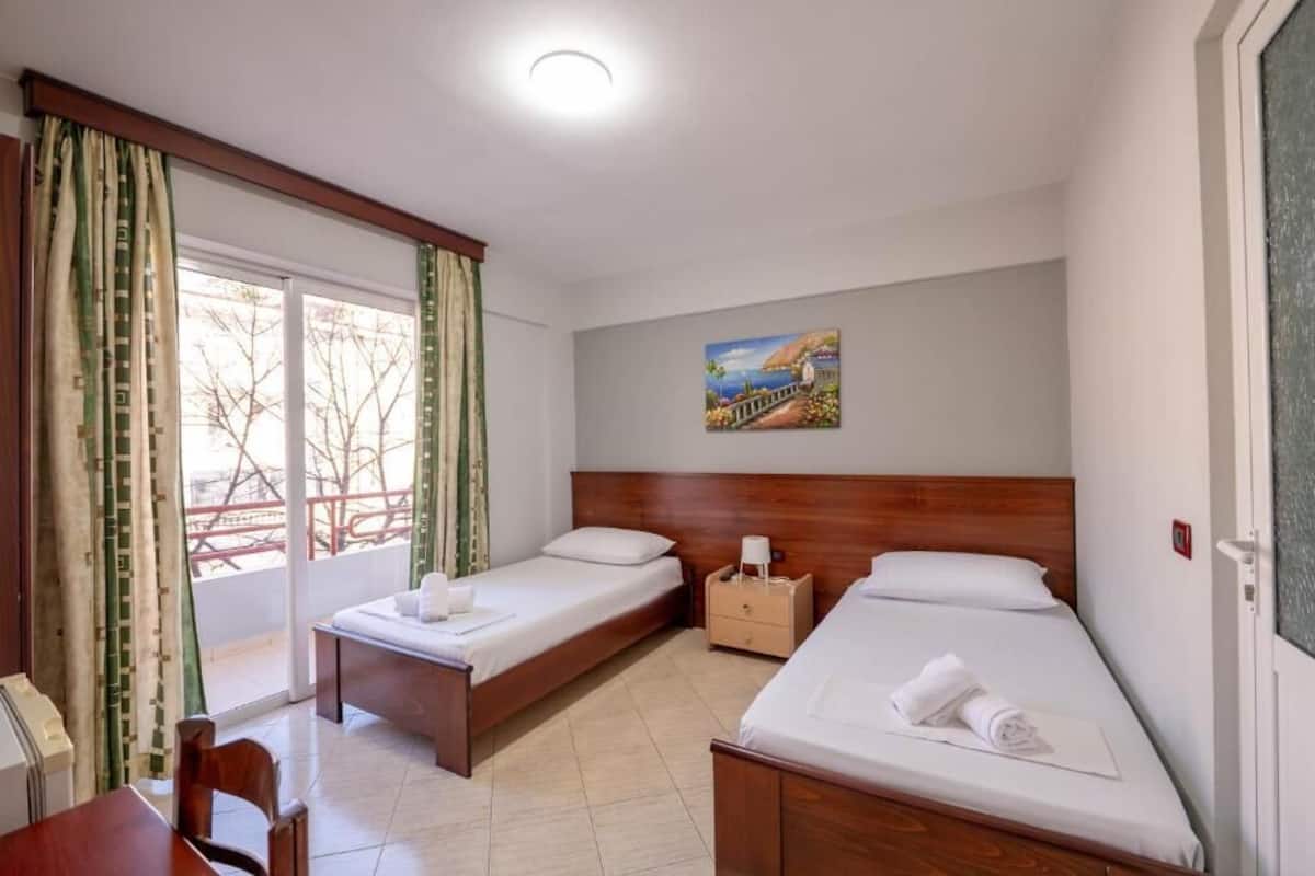 A hotel room with two single beds, a wooden headboard, a painting on the wall, and a view through the window.