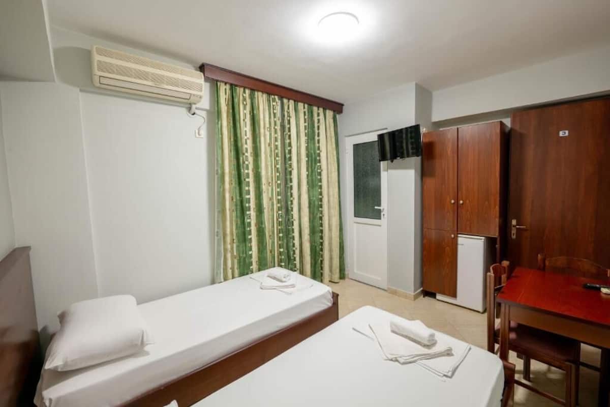 A hotel room with two single beds, a small kitchenette, a wooden wardrobe, and a dining table.