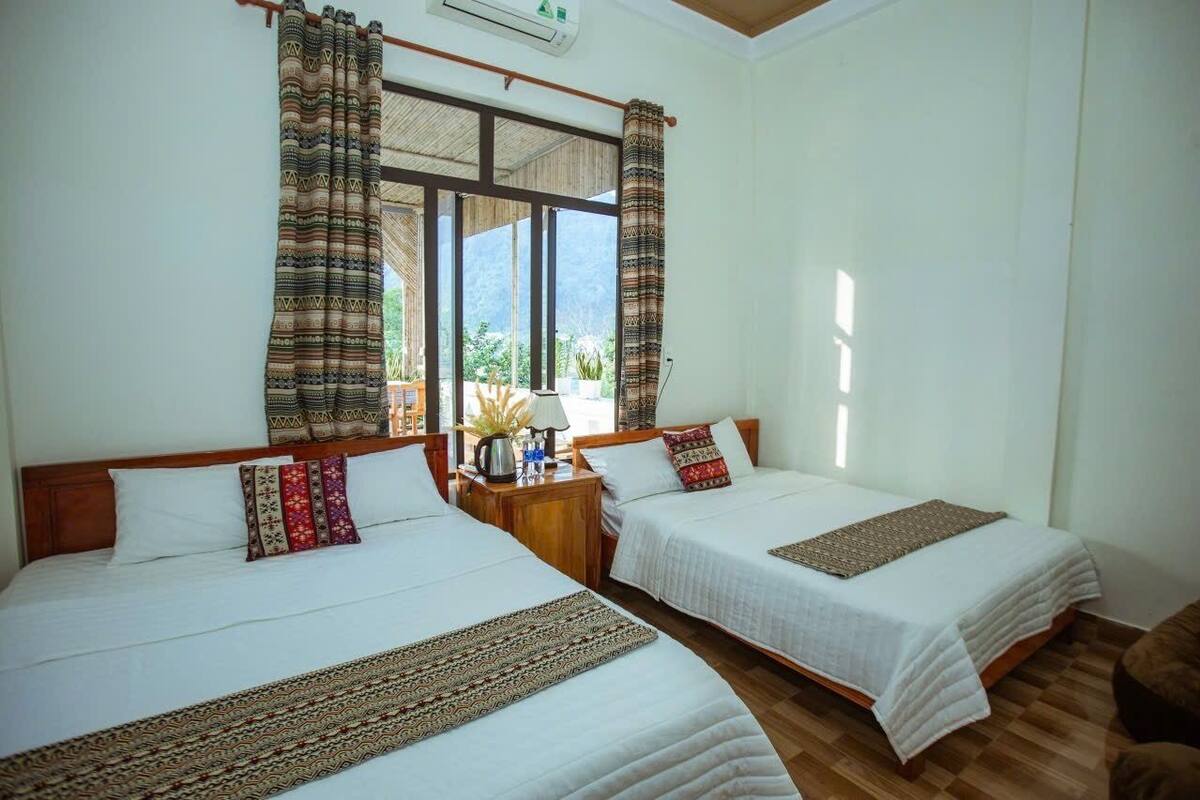 Ein Zimmer mit zwei Betten, einem Vorhangfenster, Holzboden und einem kleinen Tisch mit Lampe.