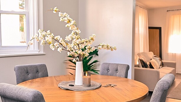 Un comedor con una mesa redonda de madera, cuatro sillas grises tapizadas y un jarrón con flores blancas.