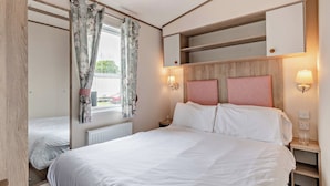 A bedroom with a bed, a window with curtains, a bedside table, and a built-in shelf above the bed. - Bronze 2 Bedroom Caravan, Sleeps 6. With parking (Great Yarmouth)