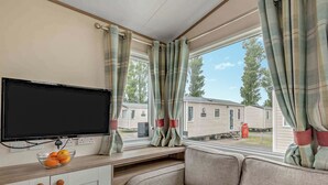 A cozy RV interior with a flat-screen TV, a bowl of fruit, and a view of mobile homes outside. - Bronze 2 Bedroom Caravan, Sleeps 6. With parking (Great Yarmouth)