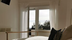 A bedroom with a wooden bed, white curtains, a view through a large window, a small round table, and a remote control on the table. - Breeze Suite with large terrace - Villa Maria (Sperlonga)