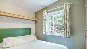 Een slaapkamer met een groen hoofdeinde, een raam met bloemen gordijnen en uitzicht op groen buiten.
