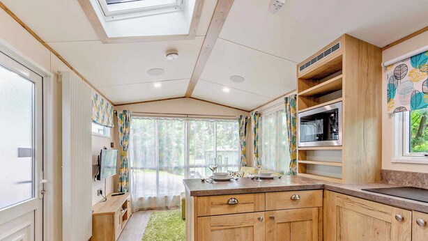 A compact kitchen with wooden cabinets, a microwave, and a dining area with a table and chairs. - Bronze Premium 1 Bedroom Caravan, Sleeps 2 (Great Yarmouth)