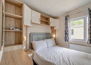 A bedroom with a bed, bedside lamps, a wooden nightstand, and built-in shelving with a pair of headphones. - Gold 3 Bedroom Caravan, Sleeps 6 (Great Yarmouth)