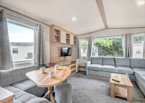 Ein gemütliches Wohnzimmer mit einer Couch, einem Holztisch mit Tassen, einem Fernseher auf einem Holzregal und großen Fenstern.