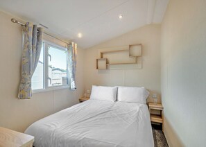 A bedroom with a bed, curtains, a bedside table, a nightstand, and a wall-mounted shelf. - Gold 4 Bedroom Caravan, Sleeps 10. With parking (Great Yarmouth)