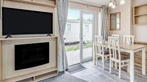 A room with a TV, fireplace, dining table, chairs, and a door leading outside. - Gold Premium Caravan with parking & ensuite (Great Yarmouth)