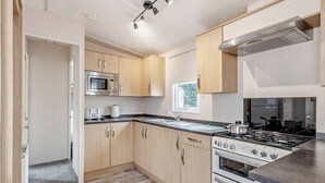 A modern kitchen with wooden cabinets, a built-in microwave, a gas stove, and a window providing natural light. - Gold Premium Caravan with parking & ensuite (Great Yarmouth)