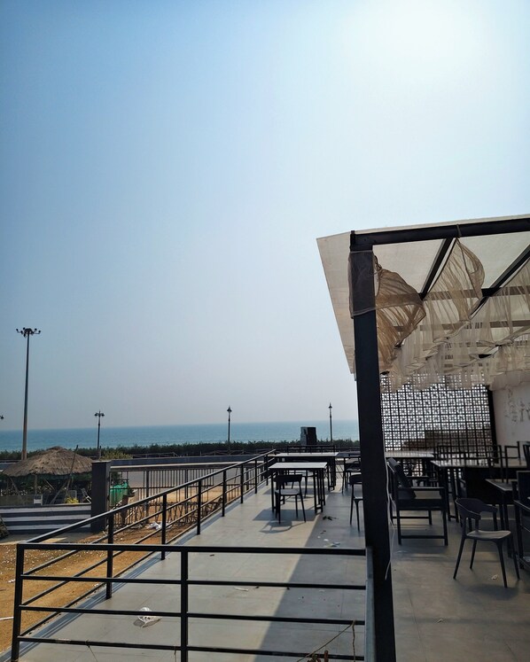 An outdoor dining area with tables and chairs, a view of the sea, and a thatched-roof structure in the background. - Hotel Bobby Beach House (Puri)