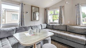 A room with a grey sectional sofa, a small table with a pitcher and cups, and a window with curtains. - Silver 3 Bedroom Caravan, Sleeps 8 (Great Yarmouth)