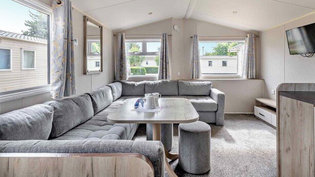 A modern RV interior with a sectional sofa, a small table, and a TV mounted on the wall. - Silver Caravan with parking (Great Yarmouth)